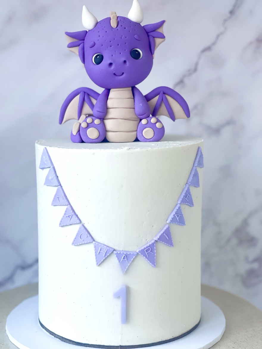 White first birthday cake with purple baby dragon topper and lavender bunting